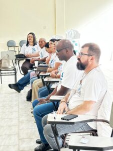Fotografia do Renato, palestrante do dia, sentado em uma das cadeiras, ao lado do Padre Derci da Silva e outros participantes do Encontro.