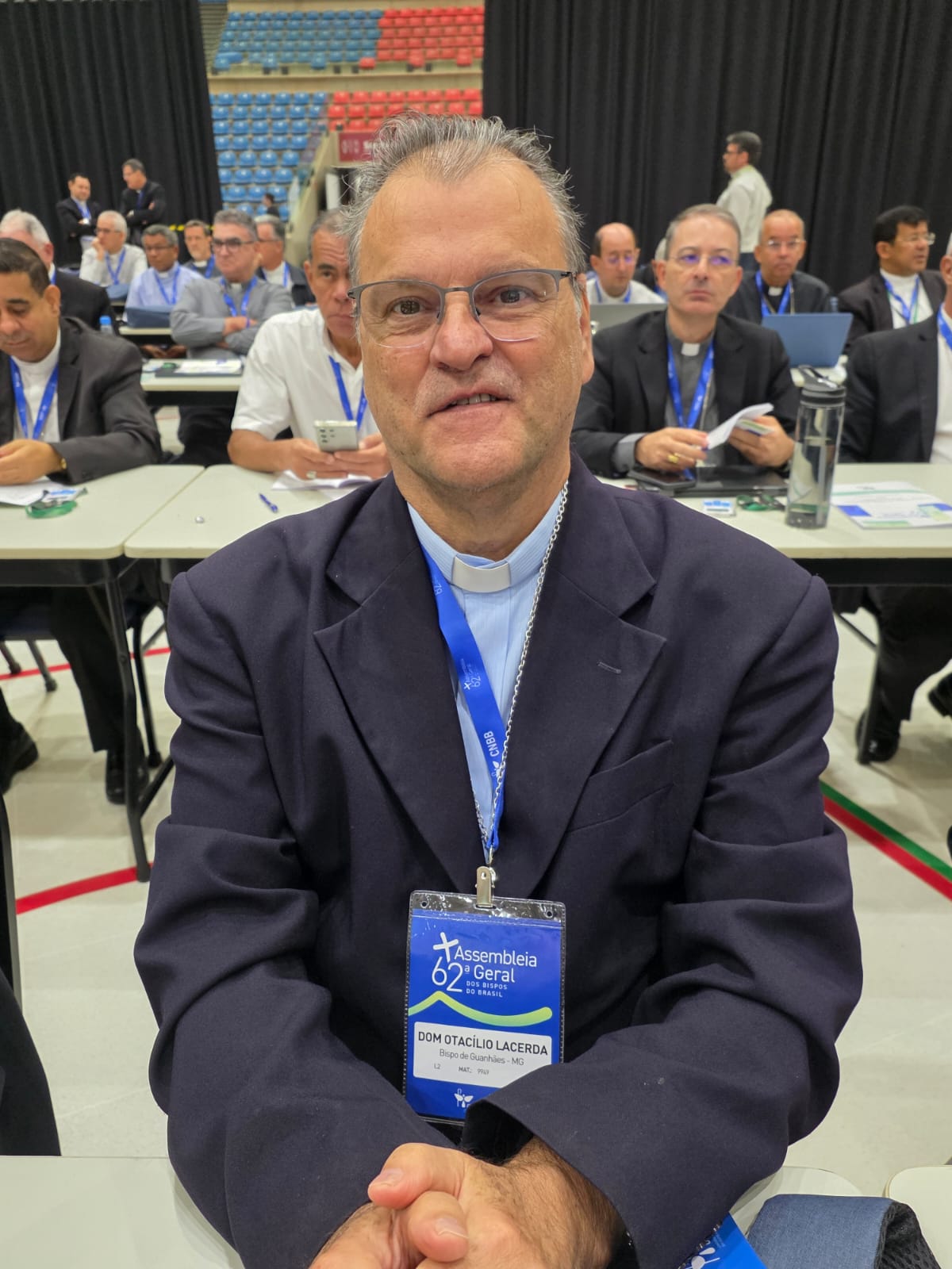 Fotografia de Dom Otacilio Ferreira de Lacerda sentado à mesa, participando da 62ª Assembleia Geral da CNBB em Aparecida. O bispo da Diocese de Guanhães está em destaque na imagem, olhando para a câmera e sorrindo. Ao fundo, outros bispos participantes da Assembleia, com leve desfoque. Na imagem é possível ver parte do local onde ocorre o evento, com cortinas longas e pretas cobrindo as arquibancadas do Centro de Eventos. As mesas dos participantes estão dispostas em grupos de fileiras laterais no centro da quadra.